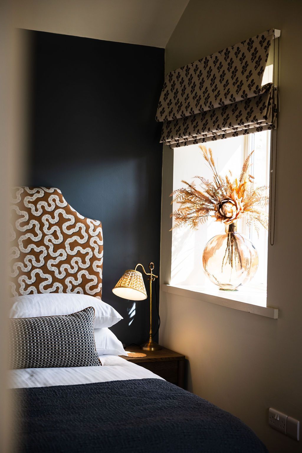 Cozy bedroom corner with dark wall, patterned pillows, bedside lamp, and a window with a floral blind.