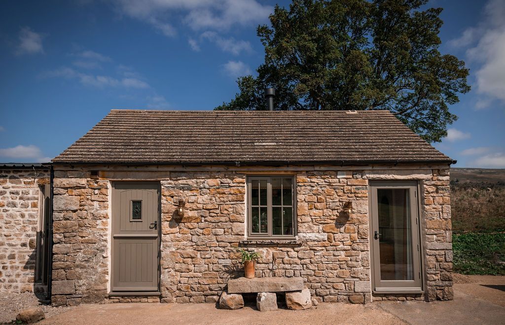 Small stone cottage with a tiled roof, wooden doors, and a tree beside it under a blue sky