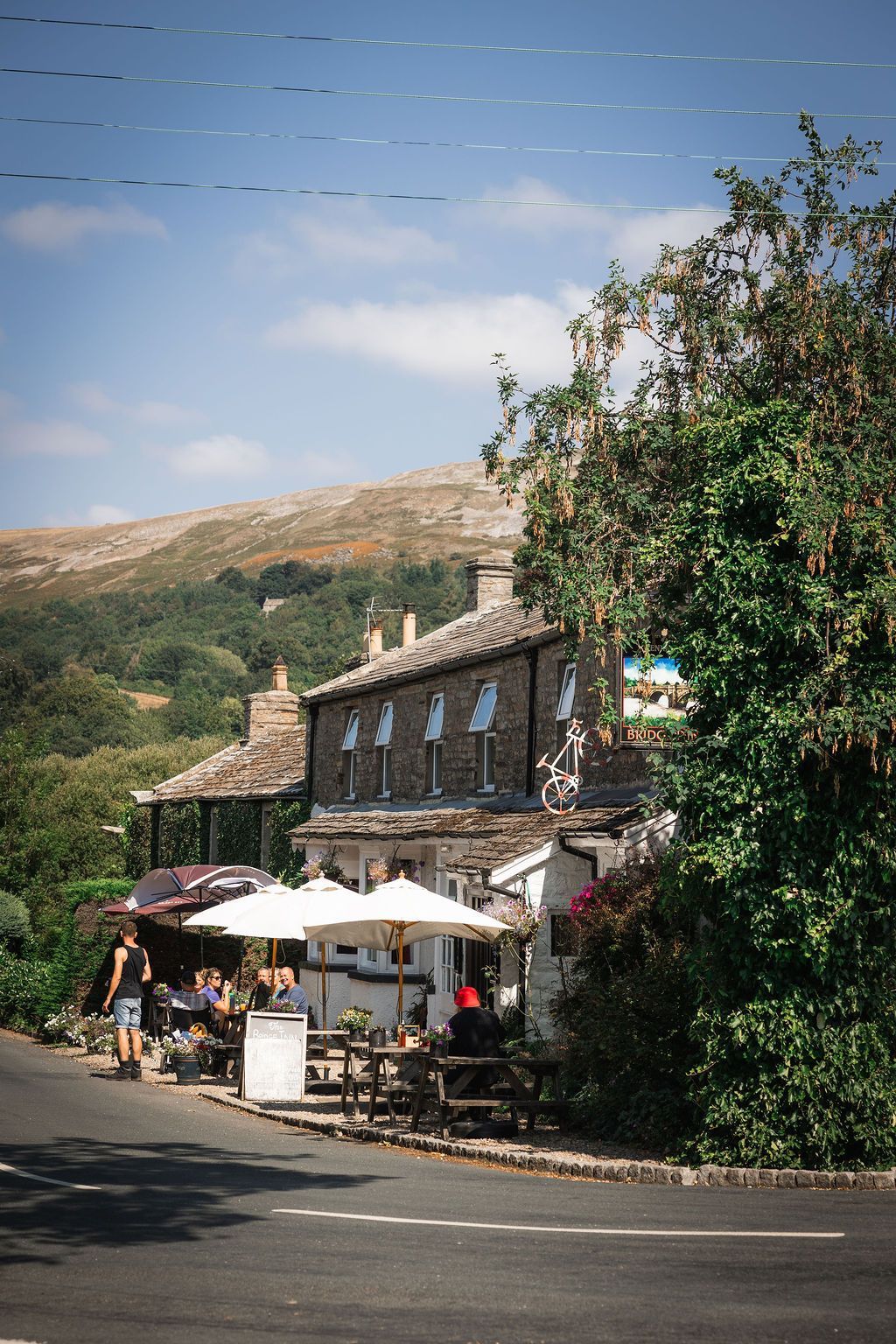 Stone building with outdoor café tables and umbrellas beside a winding road in a sunny hillside village