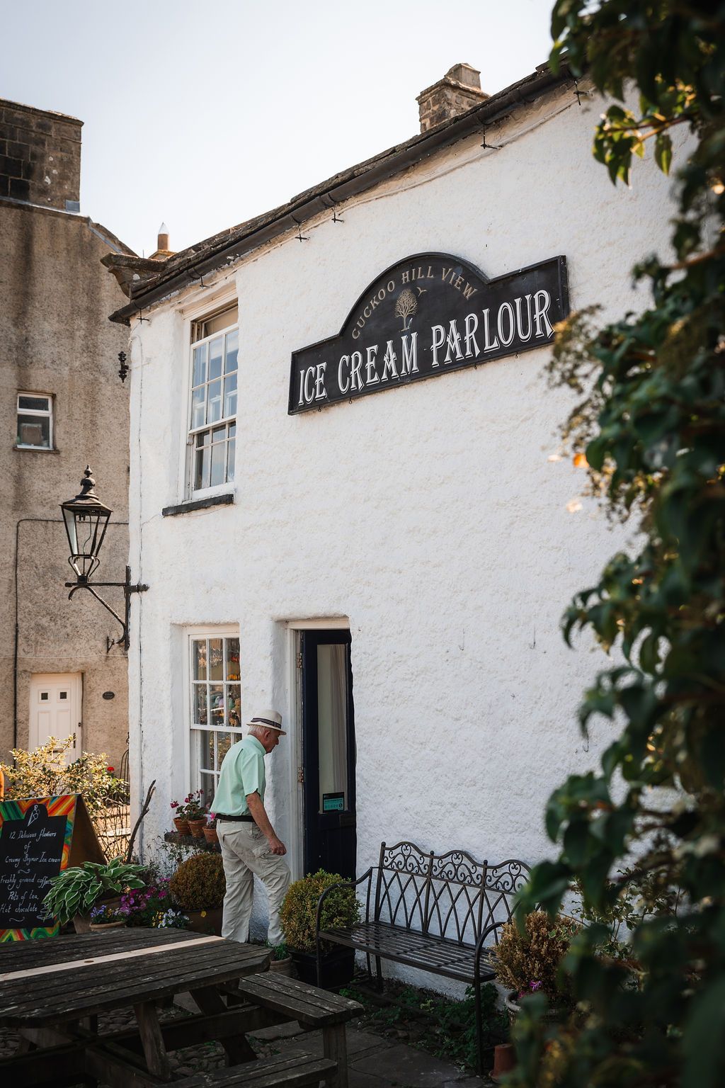 White pub exterior with black sign; a person stands in the doorway beside flower boxes and outdoor tables.
