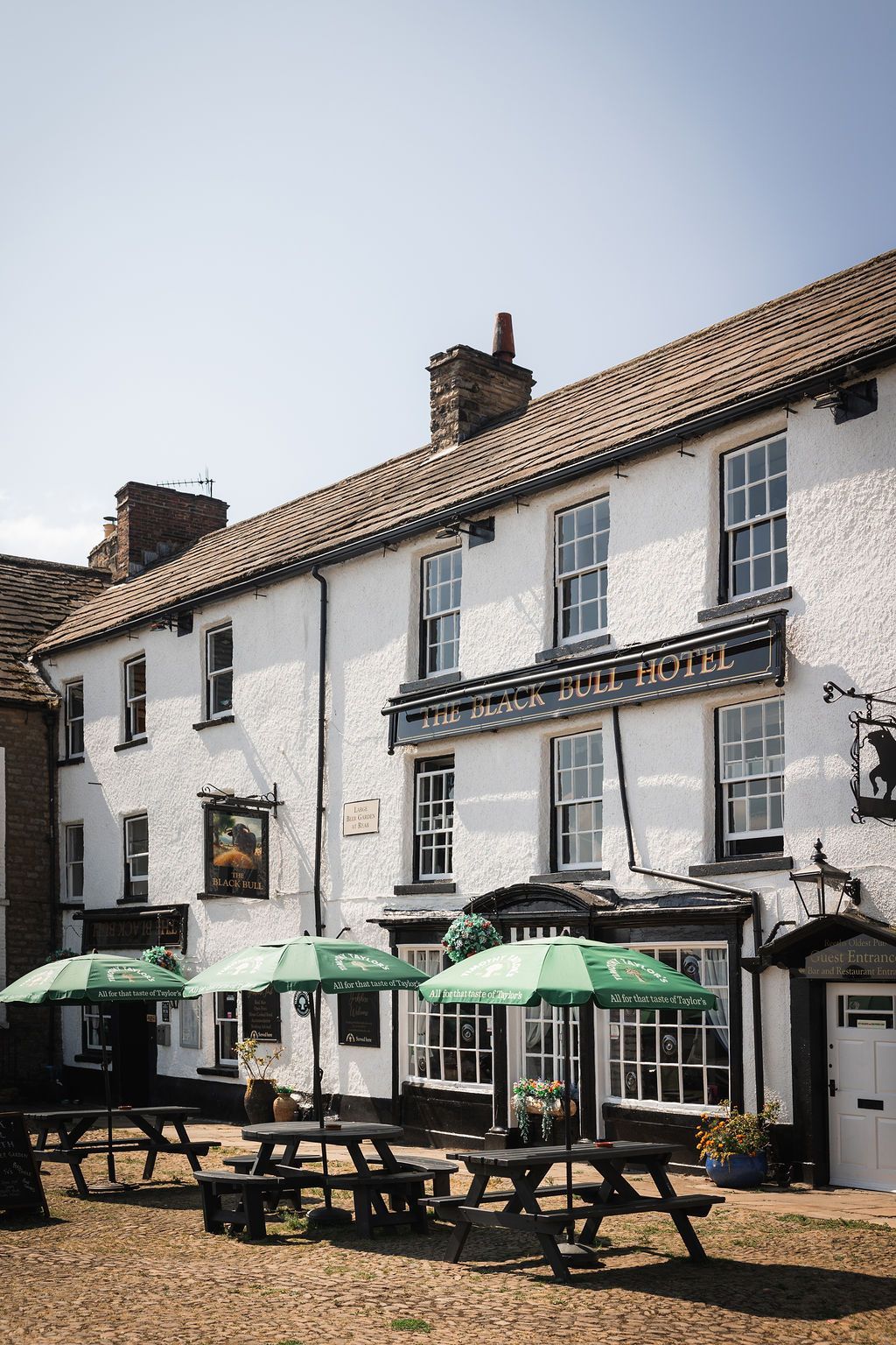 White-painted pub with green patio umbrellas and picnic tables on a sunny street.
