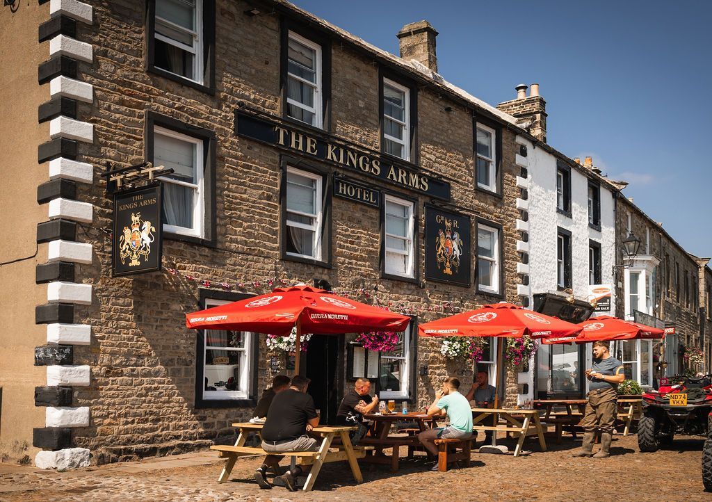 Outdoor pub with red umbrellas and people seated at picnic tables outside a brick building