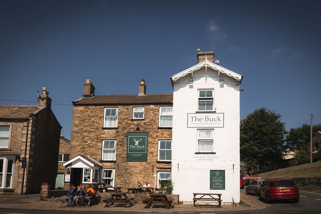 Stone pub and white building with sign in a sunny village street, with bicycles and a parked red car