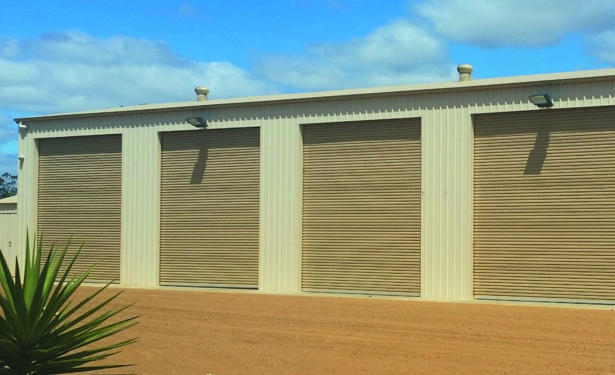 Tan building with tan roll-up doors and a white roof in a sandy yard under a blue sky