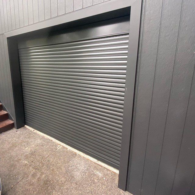 Gray roll-up garage door set in a dark-paneled exterior wall with concrete ground.