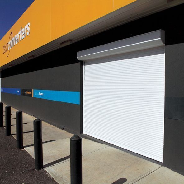 Closed storefront with white shutter, black bollards, and orange-and-black signage along a sidewalk