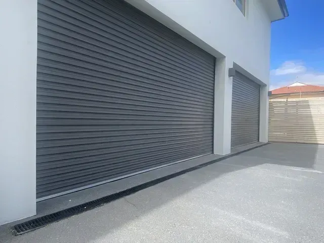 Row of gray metal roll-up garage doors along a white building beside a paved driveway.