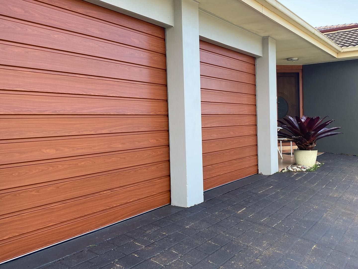 Two orange garage doors beside a white column and potted plant on a paved driveway.