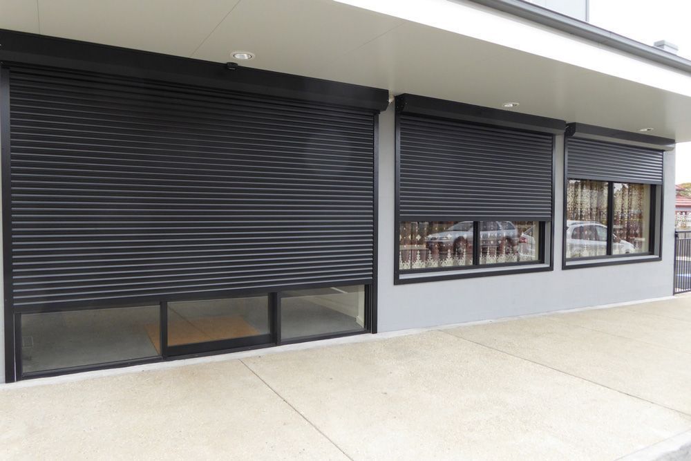 Closed black metal storefront shutters on a building exterior with windows and a concrete walkway.