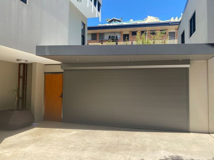 Modern building entrance with a gray garage-style door, orange side door, and concrete courtyard under blue sky