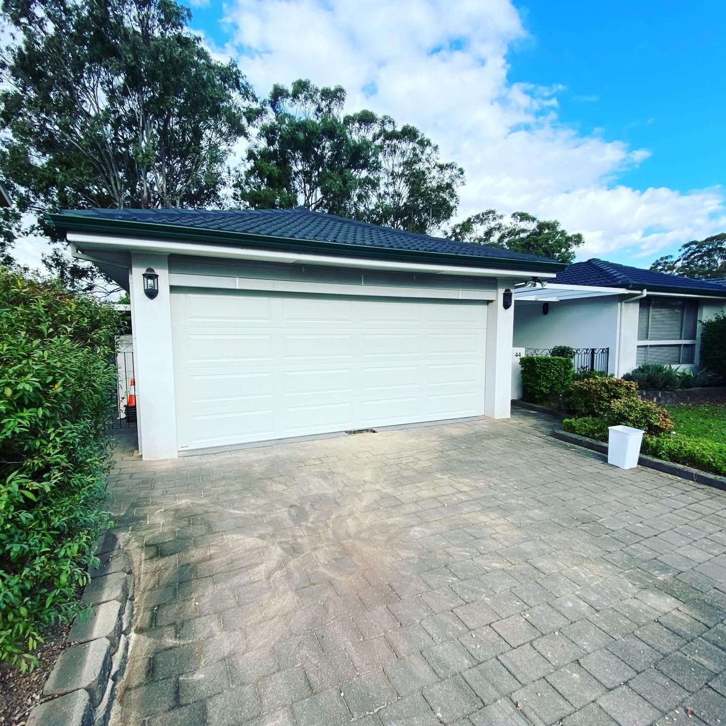 White garage with dark roof and paved driveway beside a house, surrounded by trees and greenery.