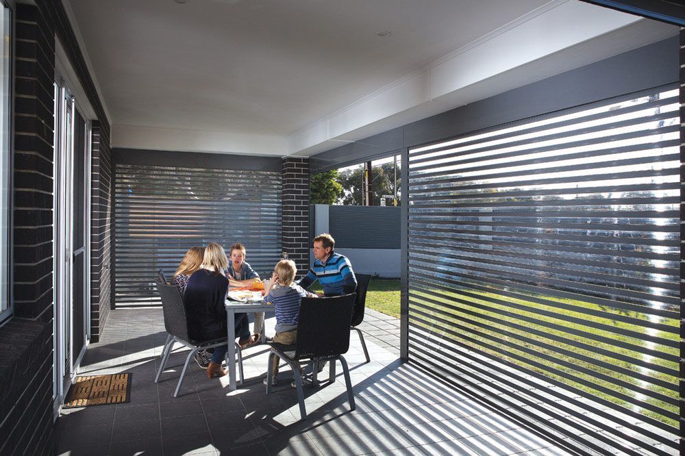 Family seated at a patio table under a shaded veranda with slatted blinds and sunlight patterns on the floor