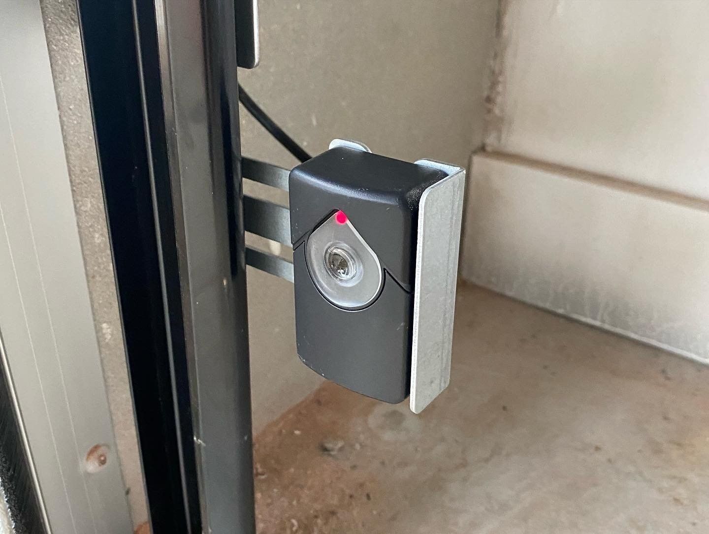 Small gray wall-mounted device with a circular lock or lens and red indicator light near a doorway.