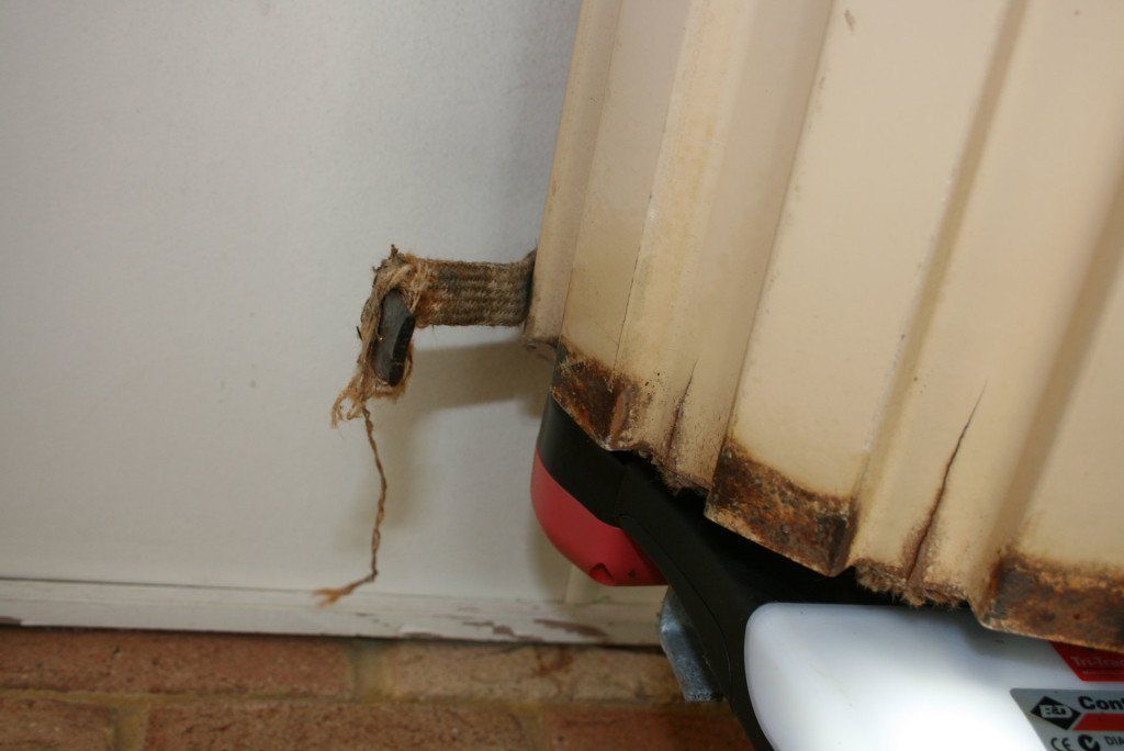 Rusty damaged pipe protruding from a wall, with corrosion staining the baseboard and floor.