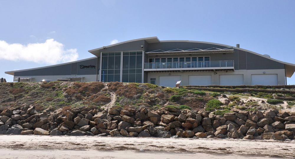 Modern building with a curved roof behind a rocky embankment and sandy beach under a blue sky