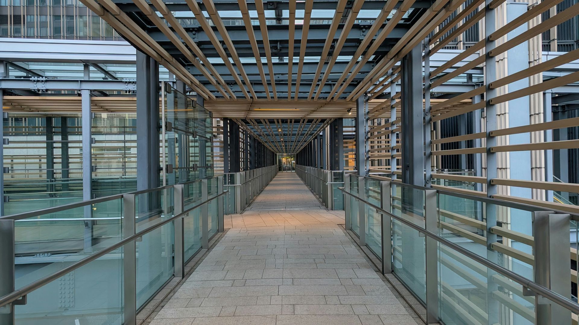 Glass-covered pedestrian walkway with metal beams and a tiled floor, seen in perspective down the center