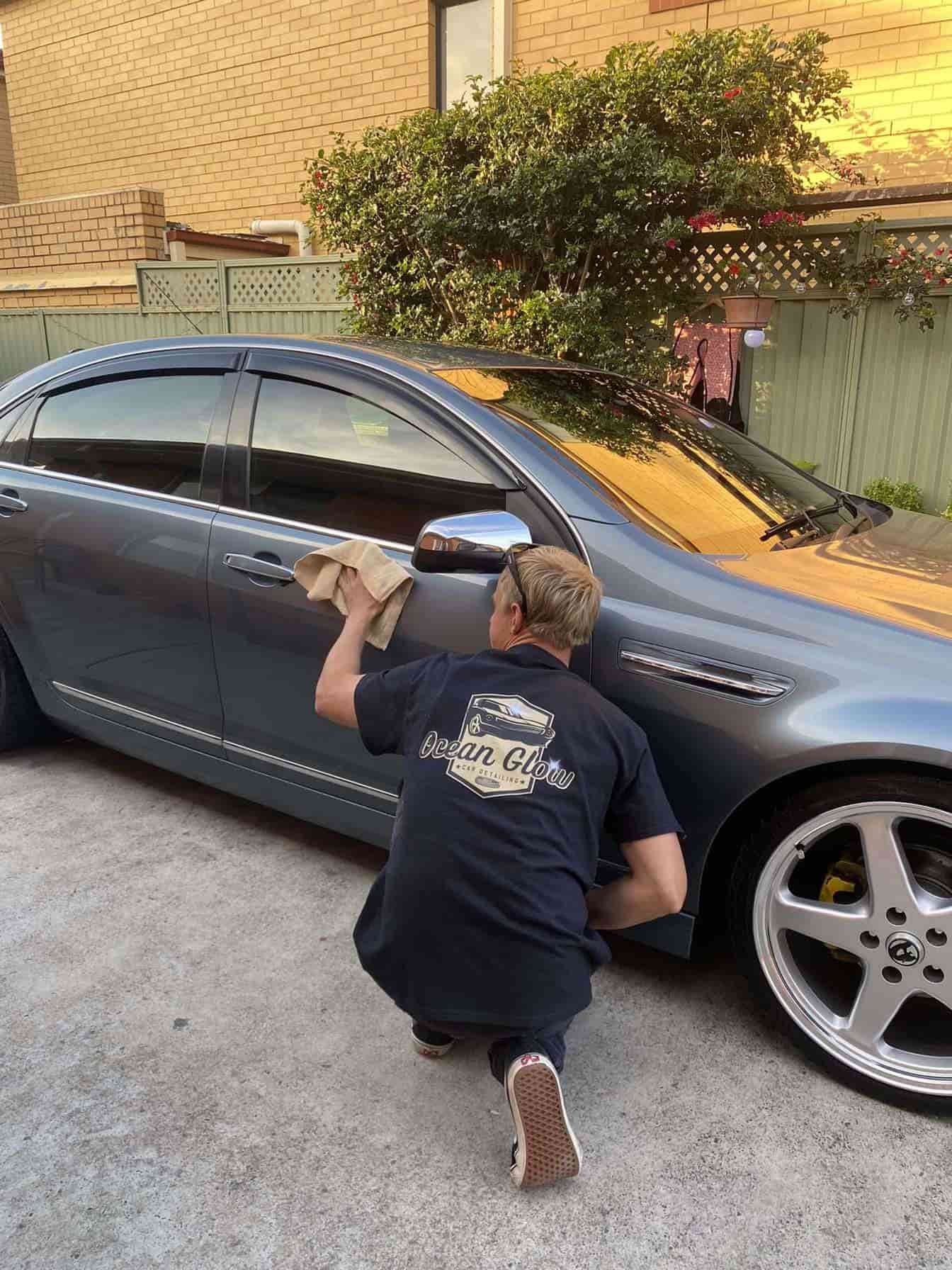 A Man Is Kneeling Down Next to A Car and Cleaning It with A Cloth – Ocean Glow Car Detailing in Long Jetty NSW