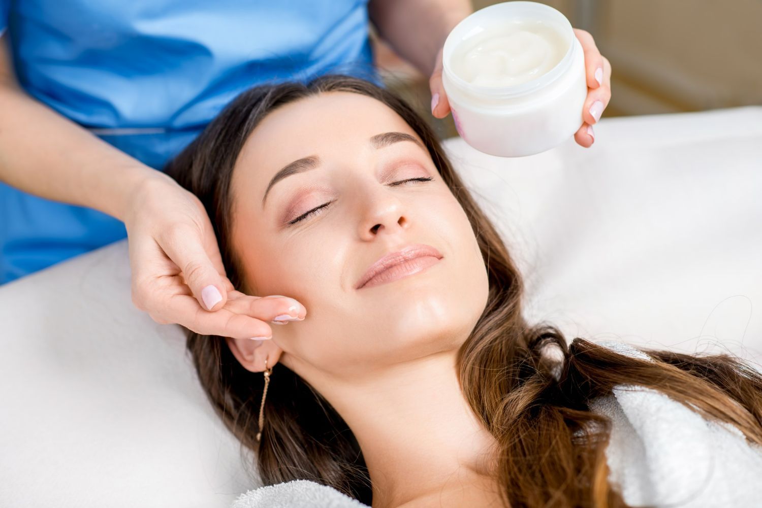 Woman receiving facial treatment; therapist holding cream jar, touching face.