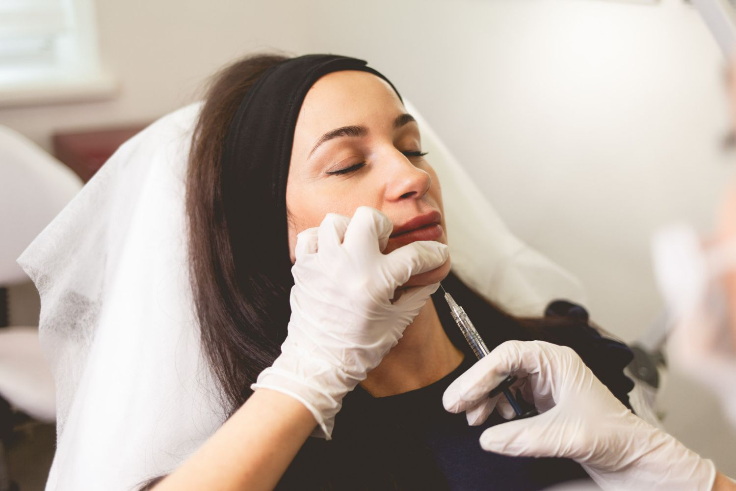 Woman receiving lip injections. White-gloved hands holding syringe near her mouth. She is wearing a black headband.
