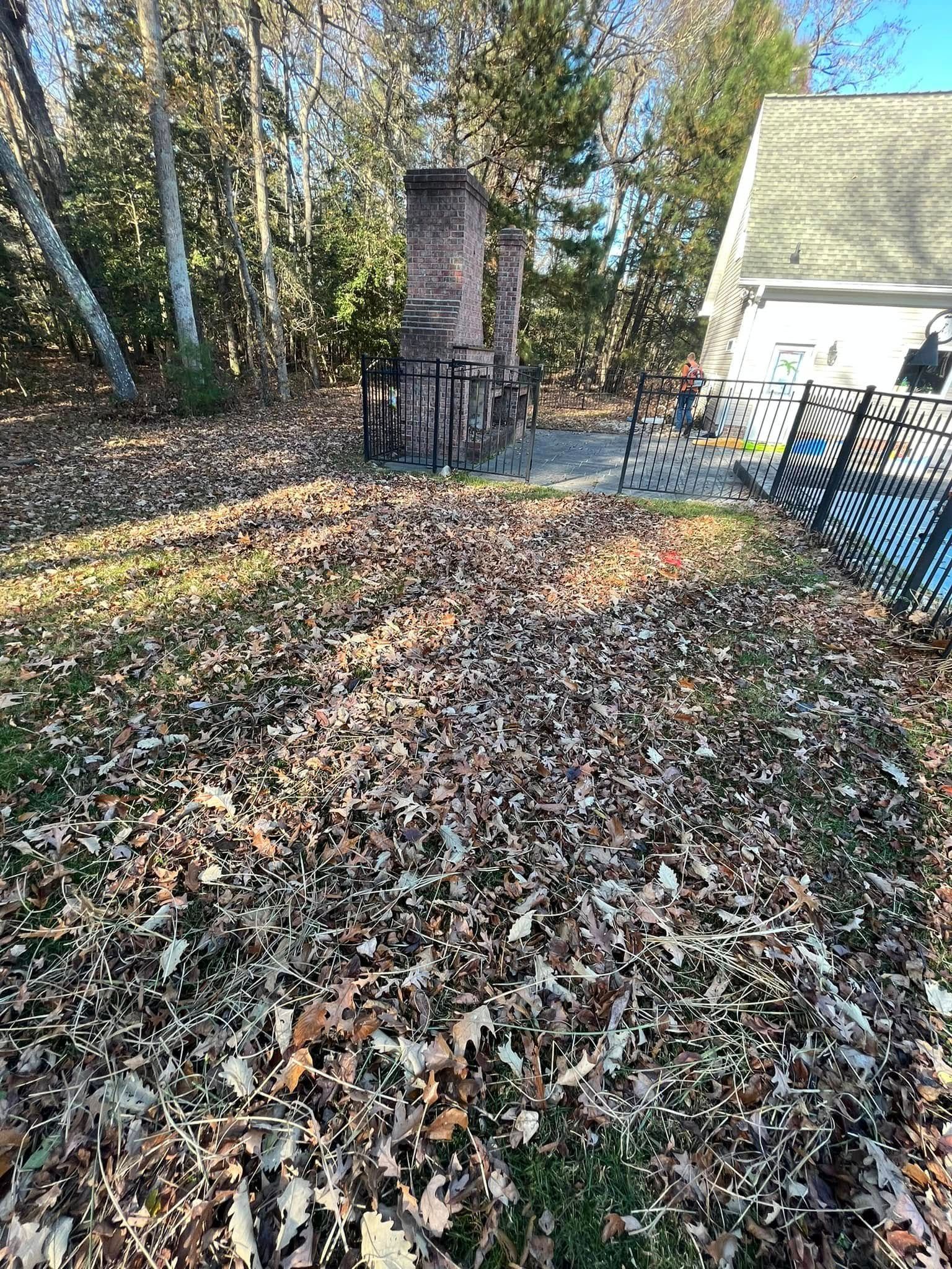 Leaf-covered yard with a brick chimney and a white house beside autumn trees.