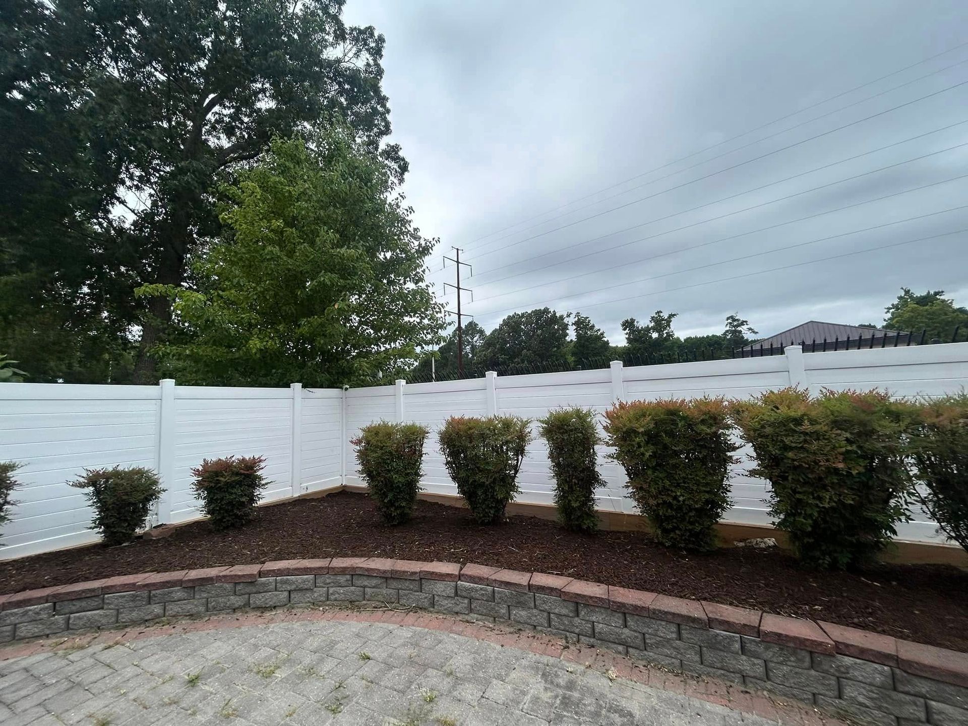 Landscaped yard with white fence, trimmed shrubs, and brick retaining wall under cloudy sky