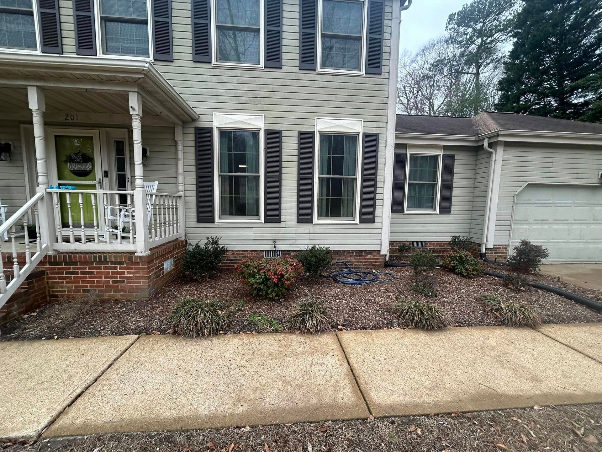 Front entrance of a beige house with white trim, porch, small garden bed, and attached garage.
