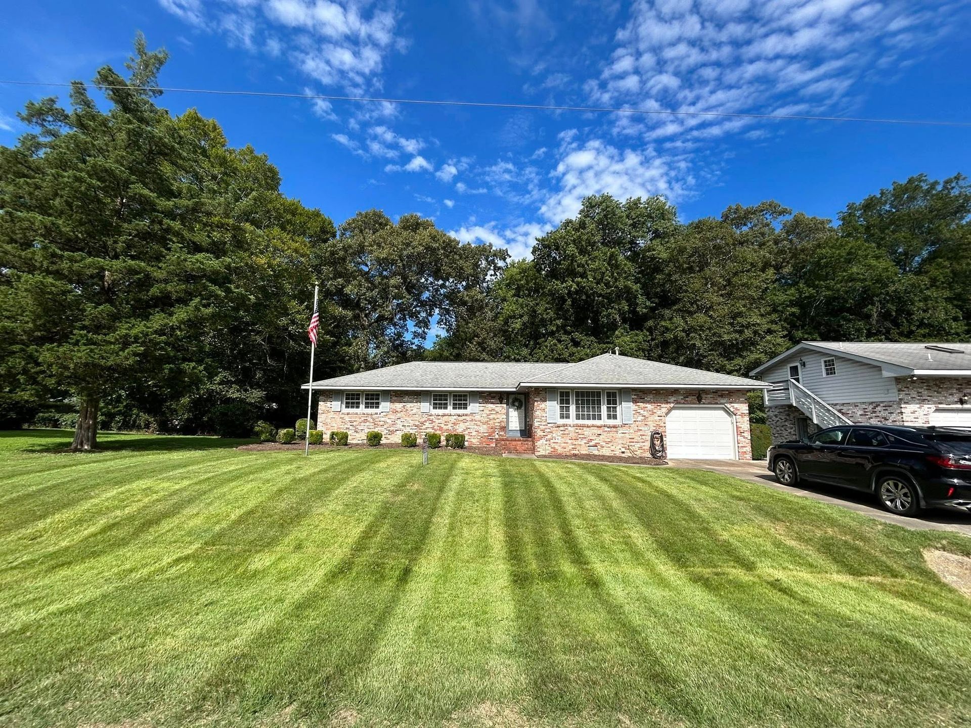 Single-story house with a green lawn, trees, blue sky, and a black car parked in the driveway.