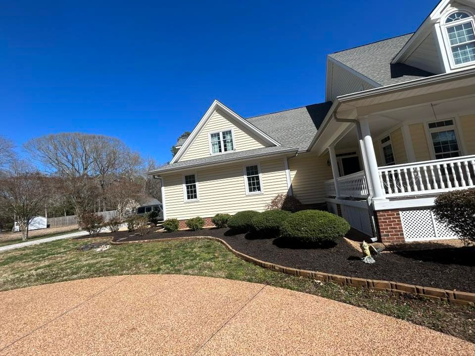 Suburban house with beige siding, white porch, manicured shrubs, and a blue sky.