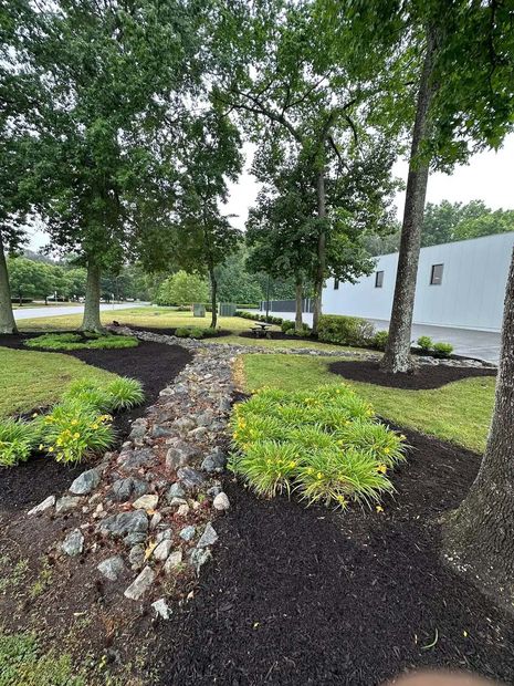 Landscaped garden with trees, mulch beds, and a rock-lined stream beside a white building.