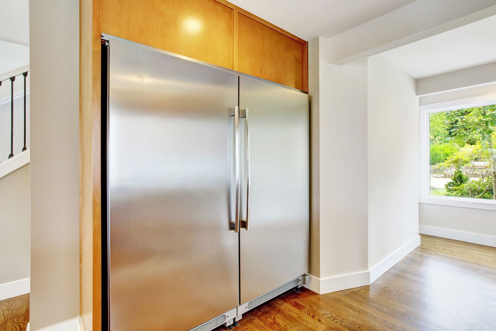 Stainless steel refrigerator built into a kitchen wall, with wooden panel above, hardwood floor, and window.