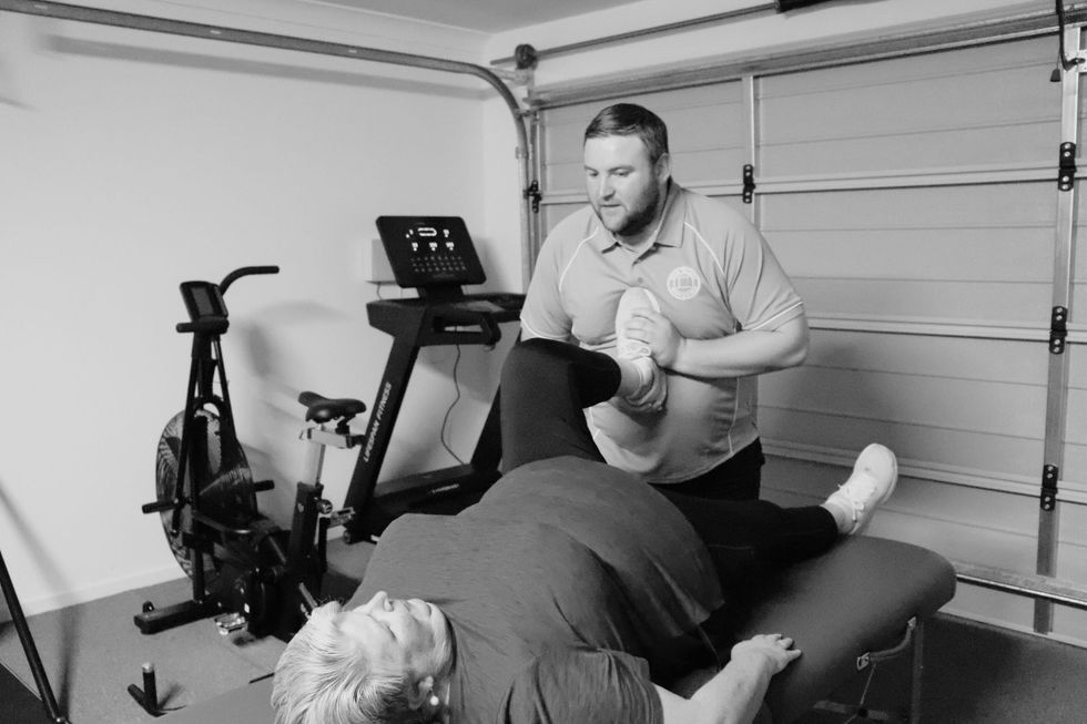 Trainer assists elderly client with leg stretch, home gym rehab.