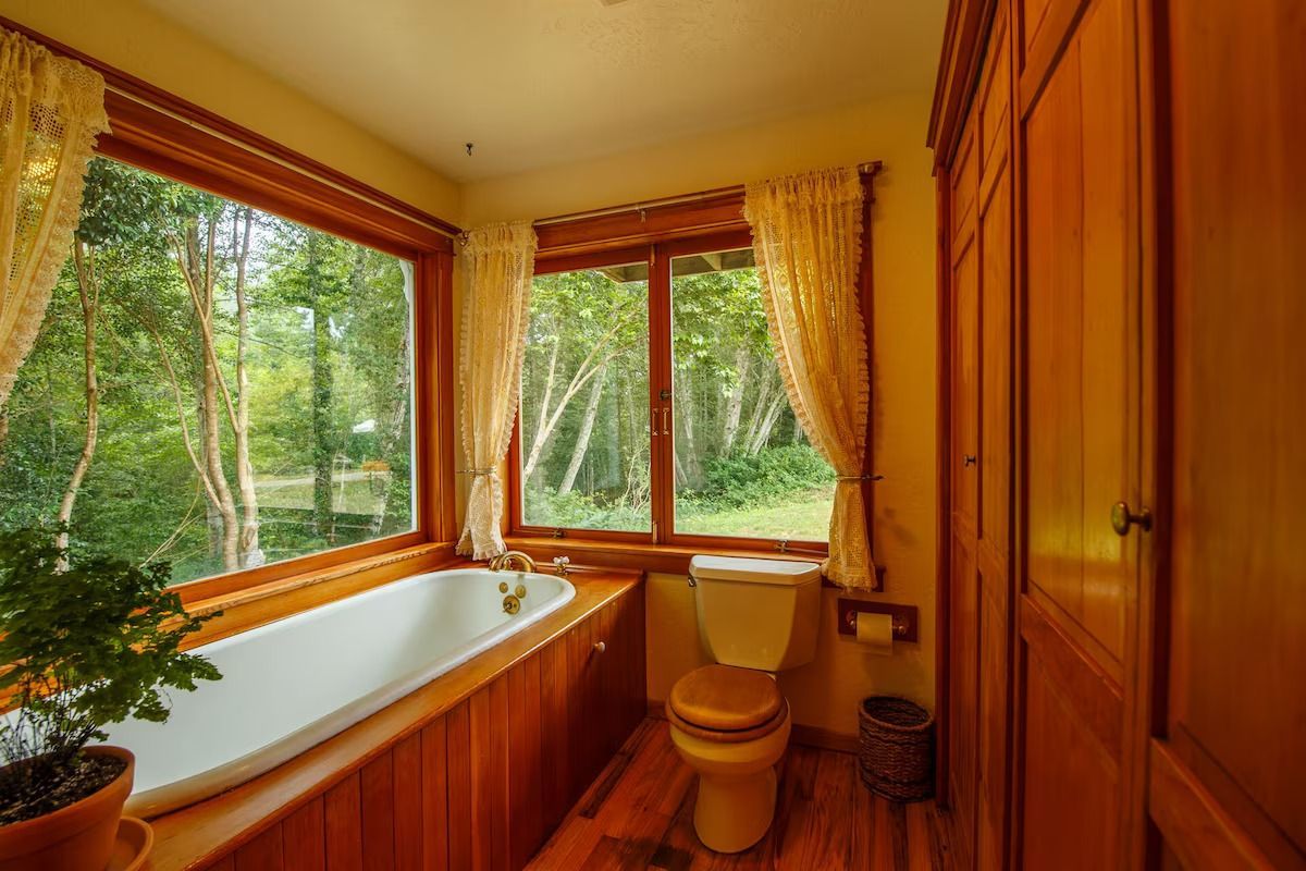 Bathroom with large windows overlooking a green landscape, featuring a bathtub, toilet, and wooden cabinetry.