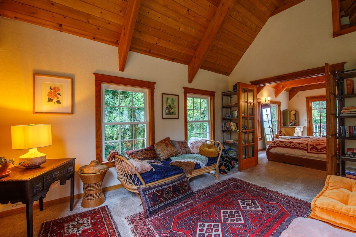 Cozy living room with patterned rug, windows, and view into bedroom. Wooden ceiling and trim.