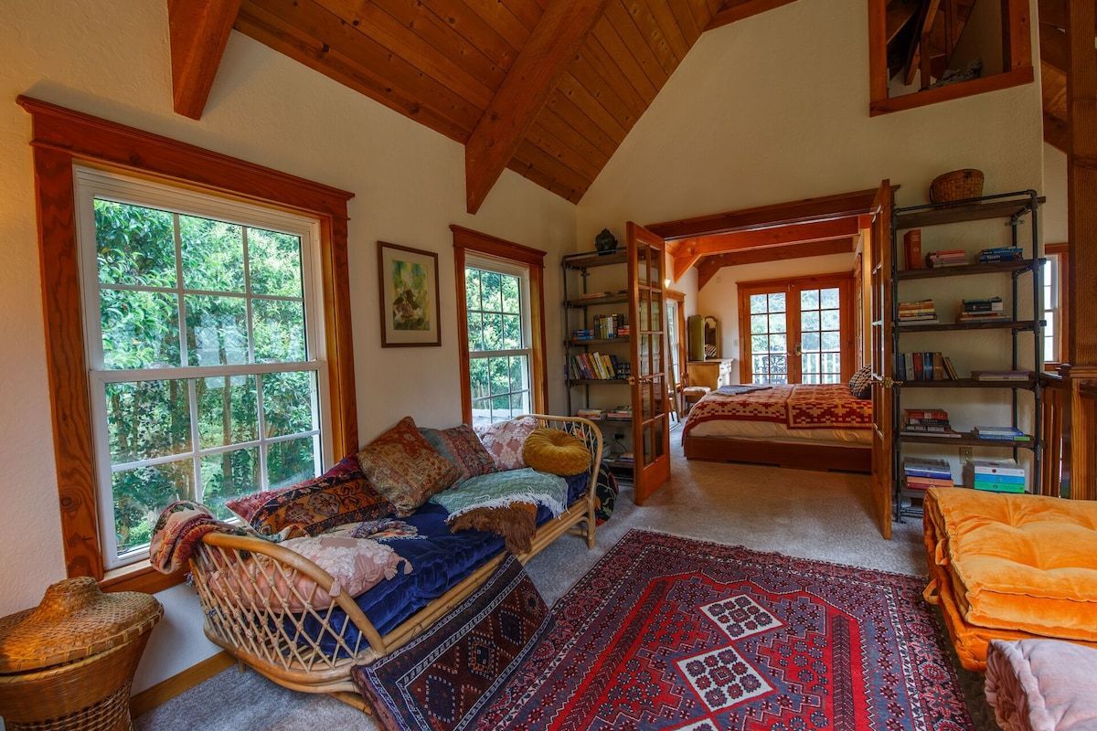 Cozy room with windows, couch, rug, and doorway to a bedroom. Sunlight streams in.
