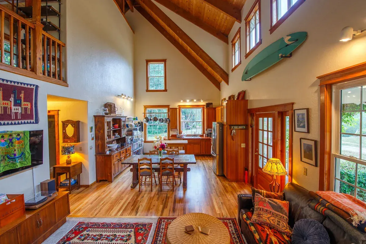 Spacious living area with high ceilings, exposed beams, and a mounted surfboard. Dining and sitting areas, hardwood floors.
