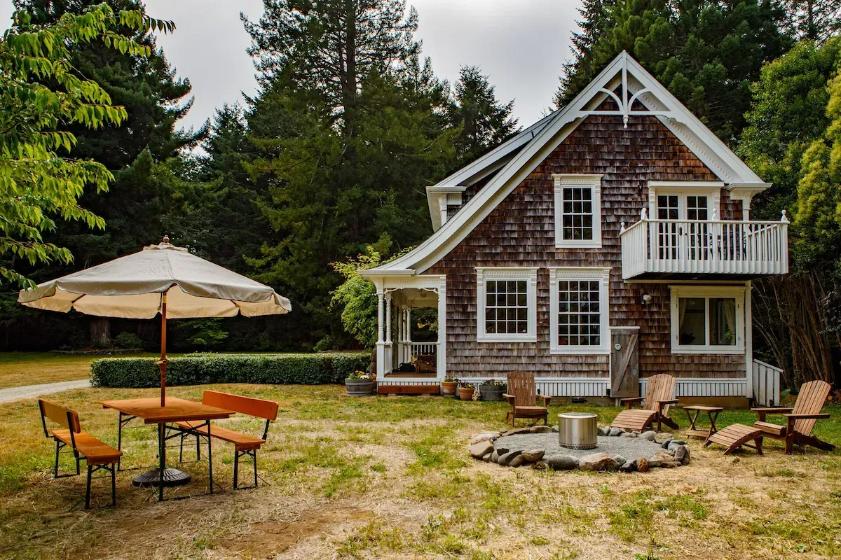 Cottage with wooden siding, white trim, and a small balcony. Yard with umbrella, table, chairs, and fire pit.