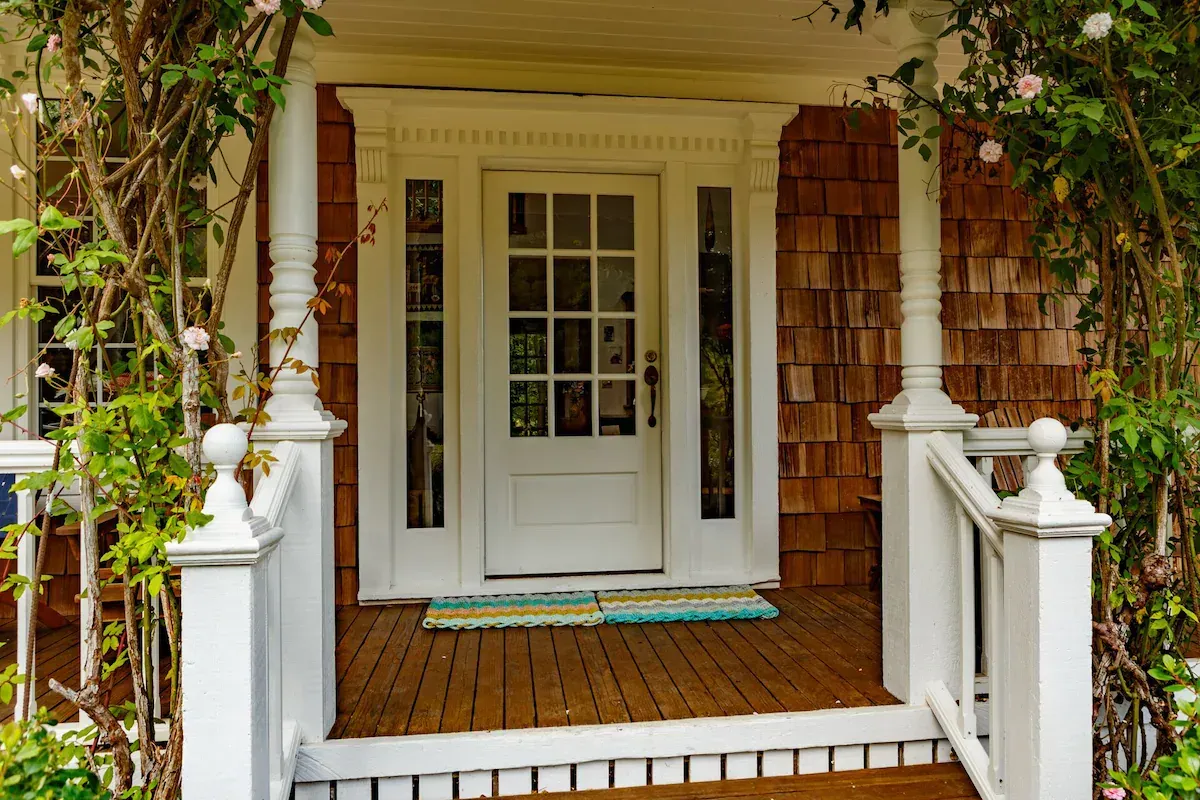 White front door with sidelights and porch, wooden shingle siding, vines and roses.