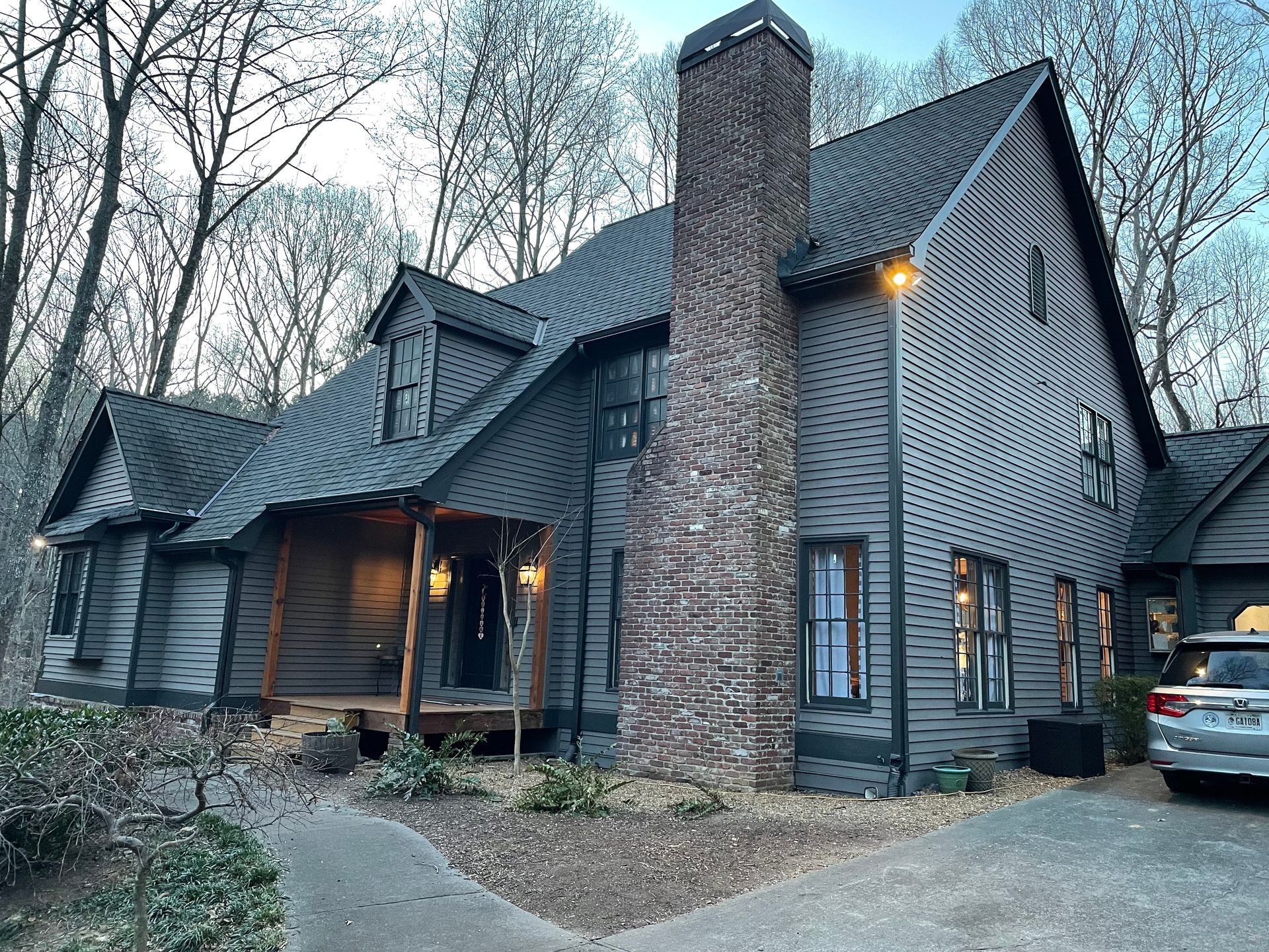 A two-story dark gray house with a stone chimney, front porch, and a car parked in the gravel driveway surrounded by trees.