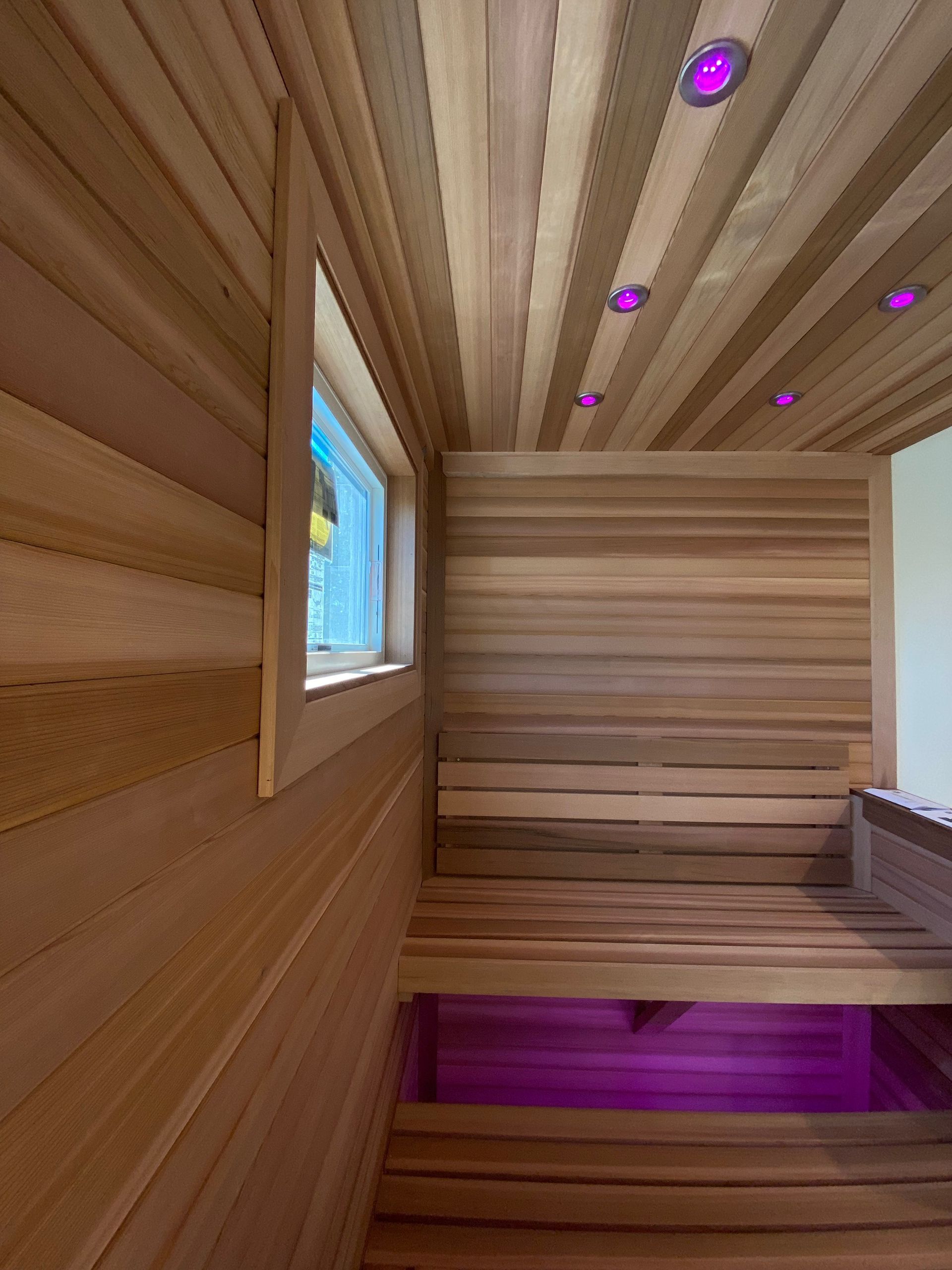 Interior of a cedar-paneled sauna featuring wooden benches and purple recessed ceiling and floor lighting.