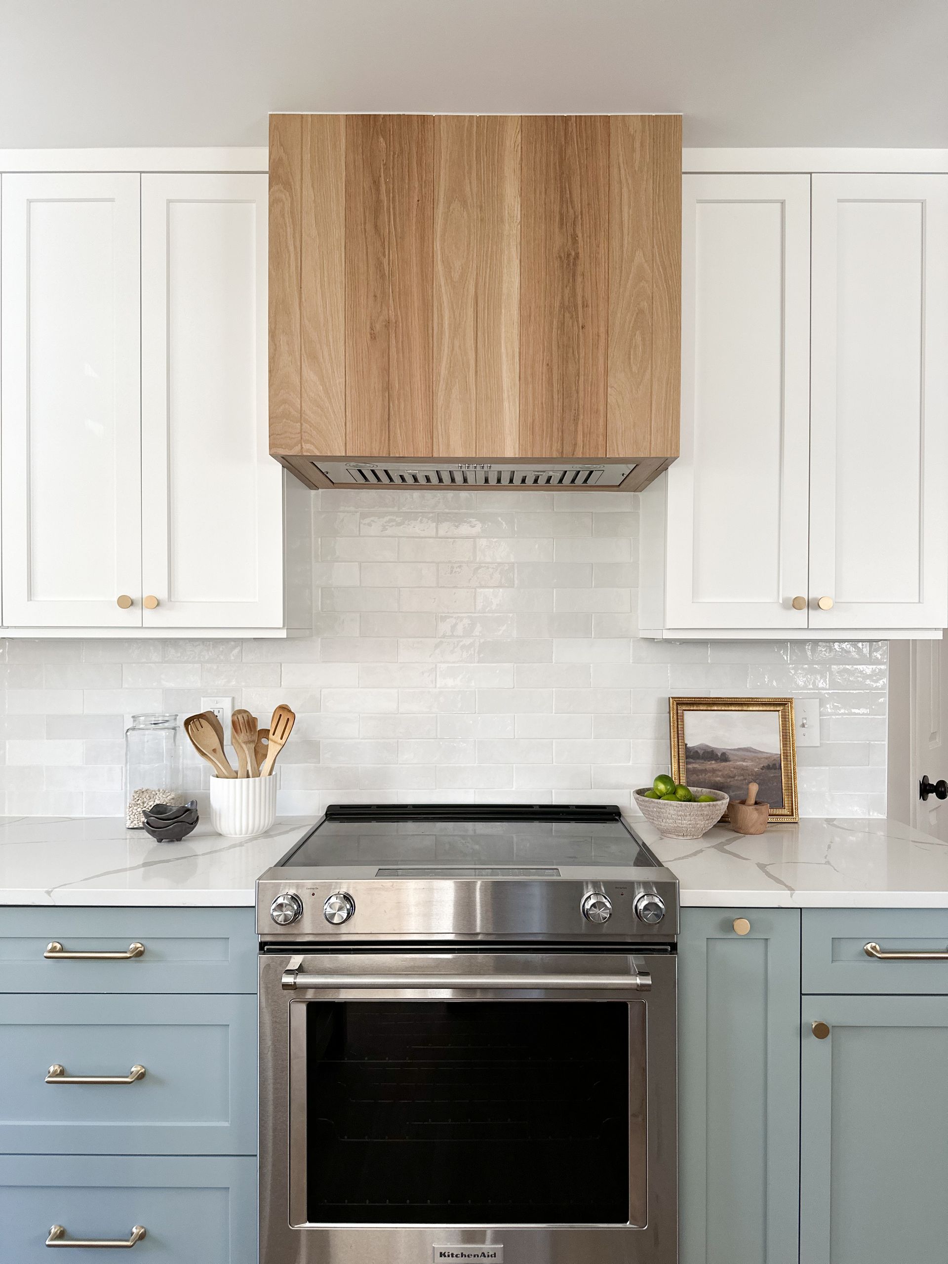 A kitchen stove with a wood-paneled range hood, flanked by white upper cabinets and light blue lower cabinets.