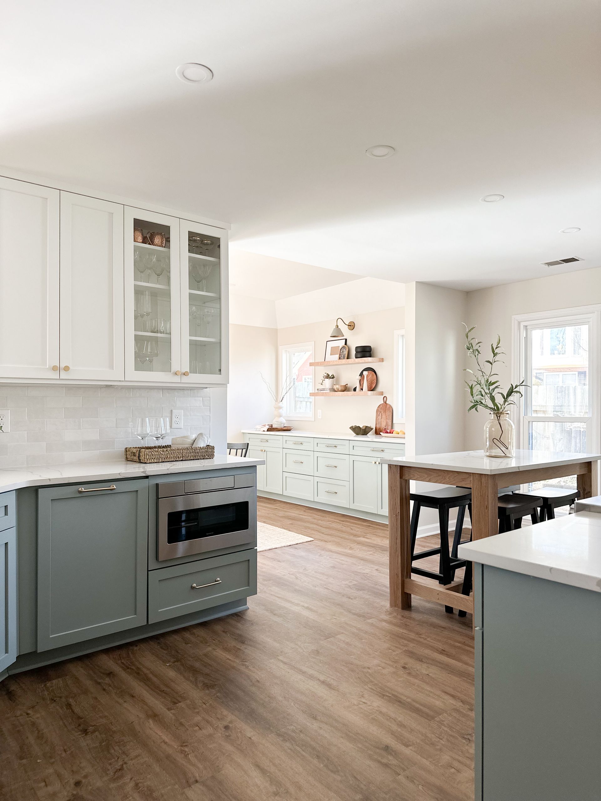 Modern kitchen with white upper cabinets, sage-green lower cabinets, wood-look flooring, and a kitchen island with stools.