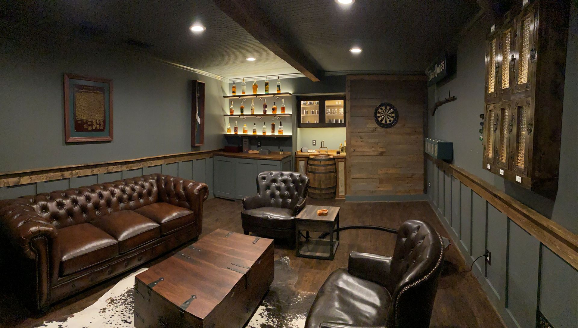 A dimly lit, wood-paneled home bar with a leather sofa, two armchairs, a rustic coffee table, and an illuminated shelf.