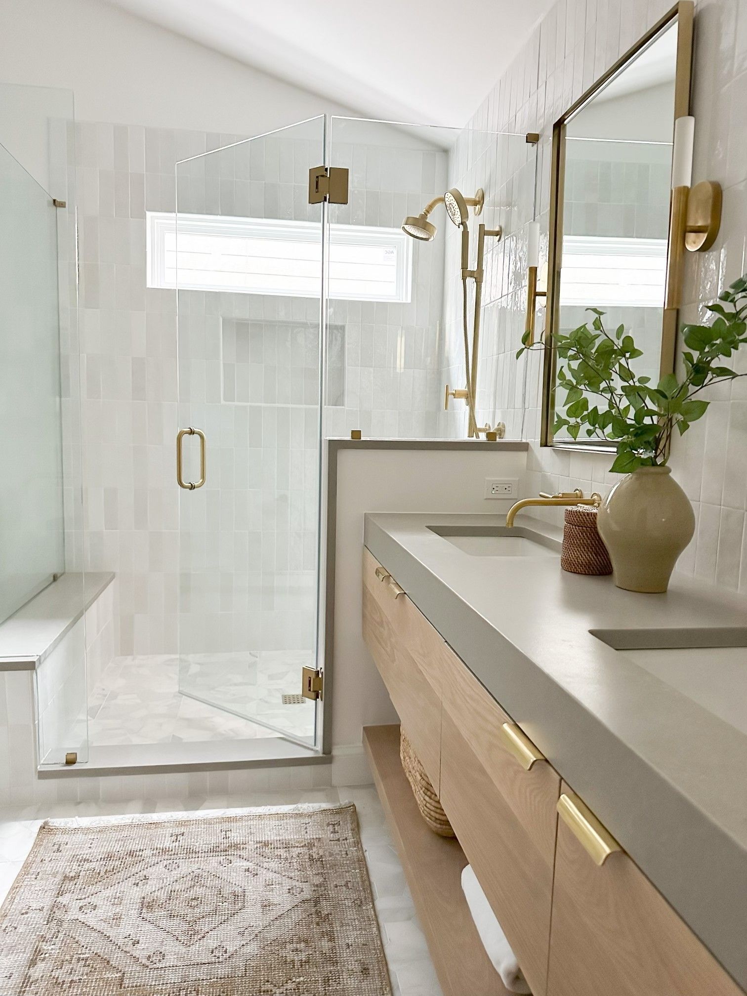 A modern bathroom featuring a light oak vanity, gray countertop, gold fixtures, and a glass-enclosed shower with white tiles.