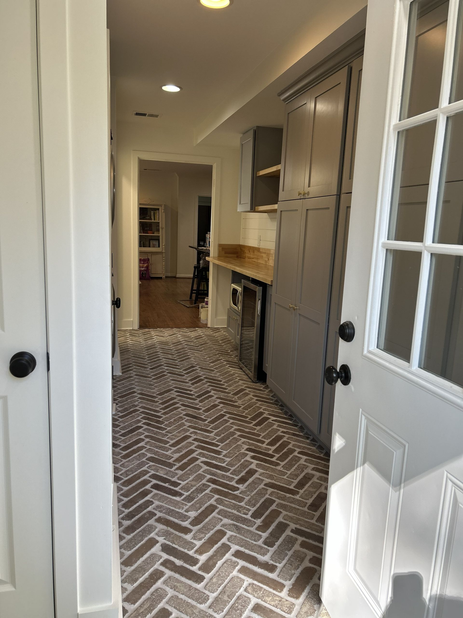 A hallway with a herringbone-patterned brick floor leads to an open room, featuring grey cabinets and a small counter.