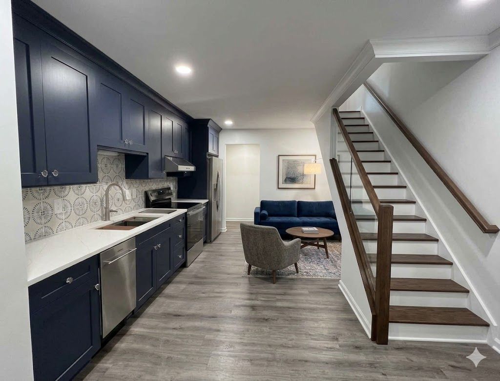 A modern, open-plan space featuring a navy blue kitchen, a living area with a blue sofa, and wooden stairs with a rail.