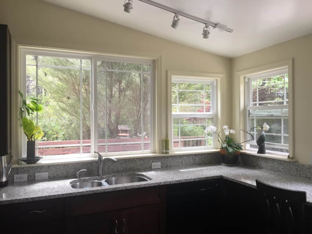 Modern kitchen with large windows, granite counters, and a sink overlooking a leafy backyard