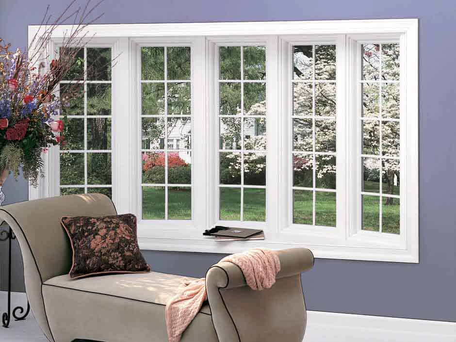 Bright bay window with white trim in a lavender room, beside a beige chaise lounge and floral pillow.