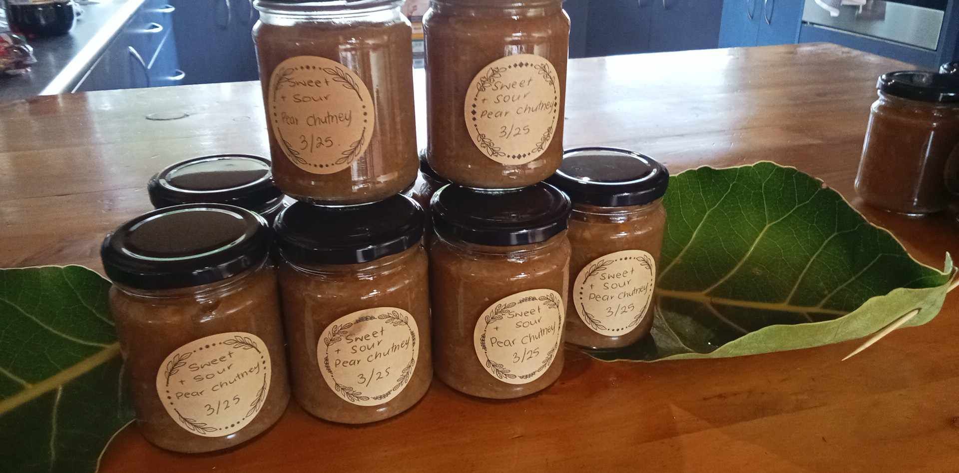 Jars of brown food, with labels, stacked on a wooden surface, with a large green leaf underneath.