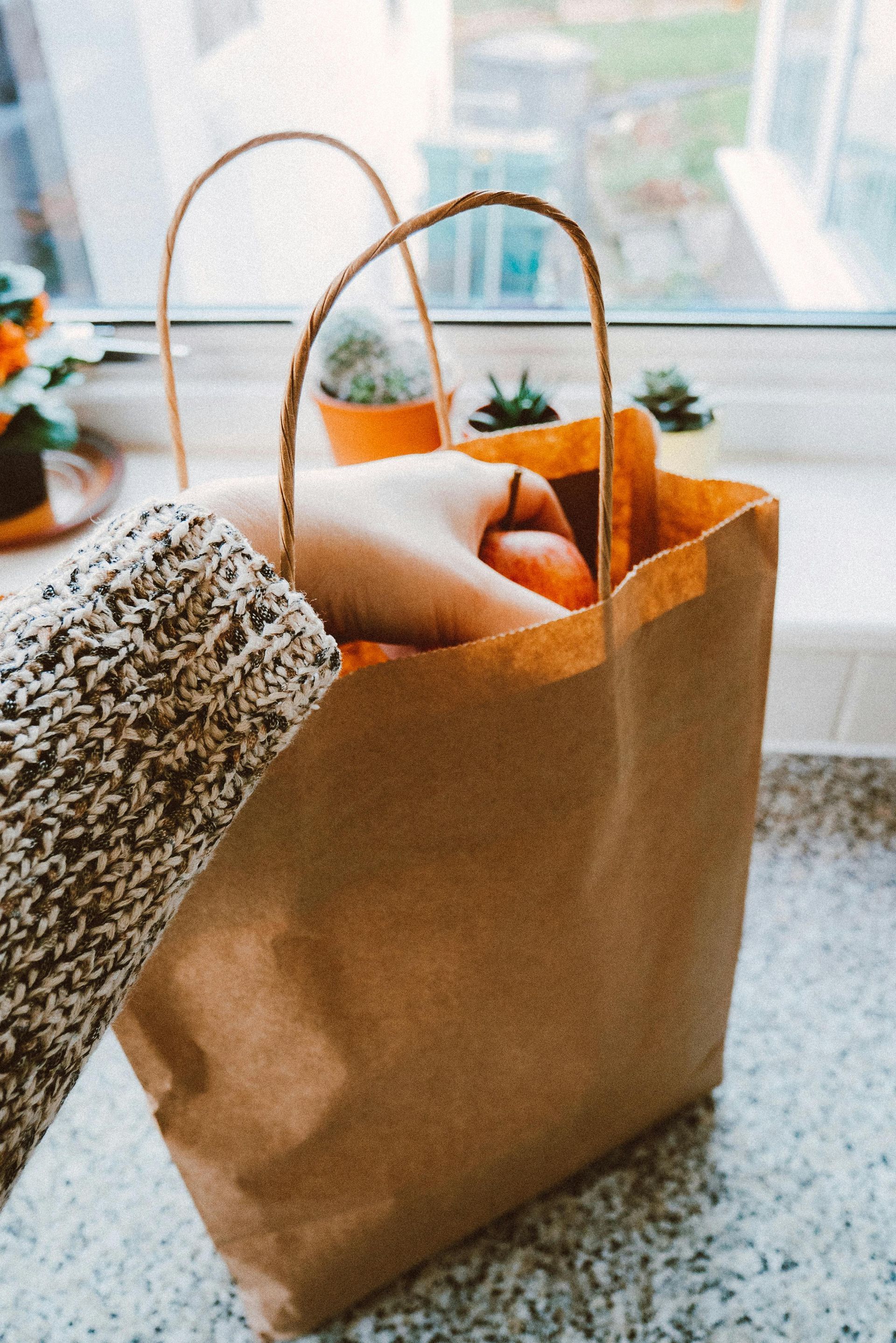 A person's hand reaching into a brown paper bag filled with oranges, placed on a countertop by a window.