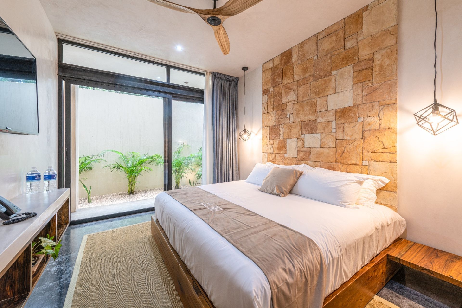 Bright modern bedroom with a bed, stone accent wall, hanging lights, and large window to a small garden